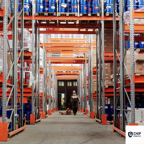 Person walking through a warehouse with high shelves stocked with blue containers and cardboard boxes.
